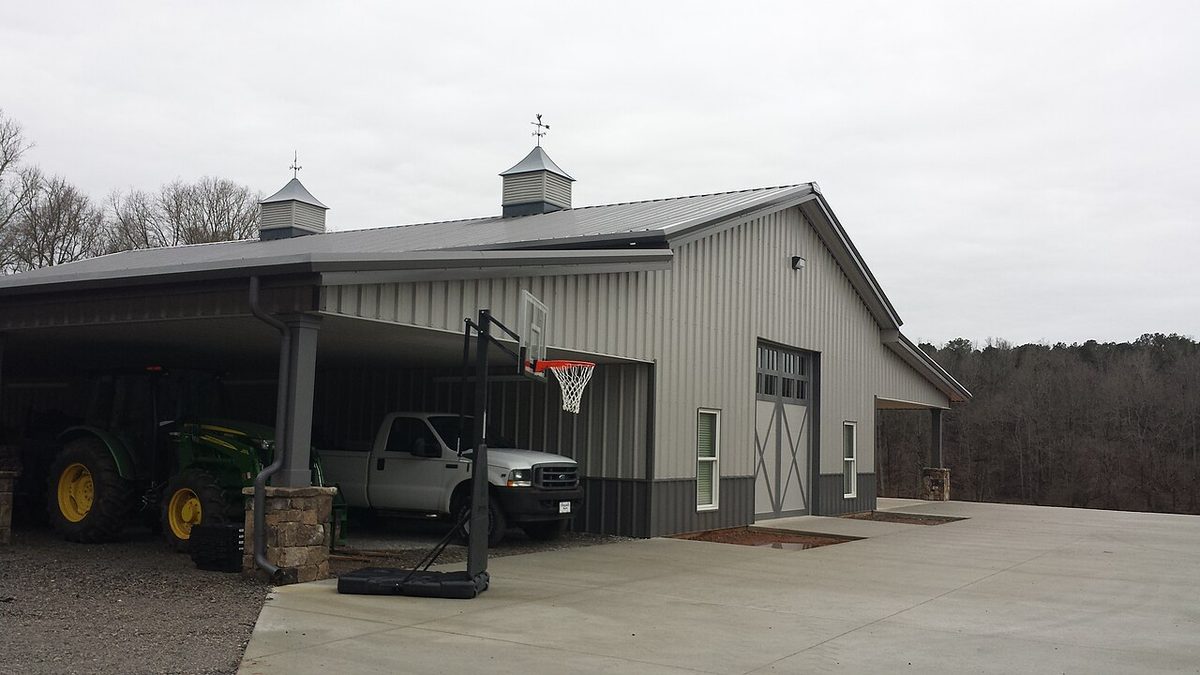 Light-and-charcoal metal shop building with cupolas, stone column bases, and equipment under a carport.